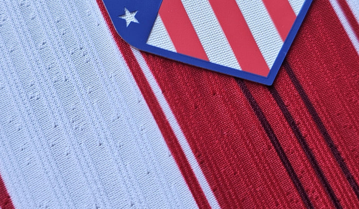 Close-up of an Atletico Madrid football shirt from the fanshop match tier. A heat-applied crest sits raised on the surface and the white stripe is a perforated engineered knit, the construction details that separate the tiers.