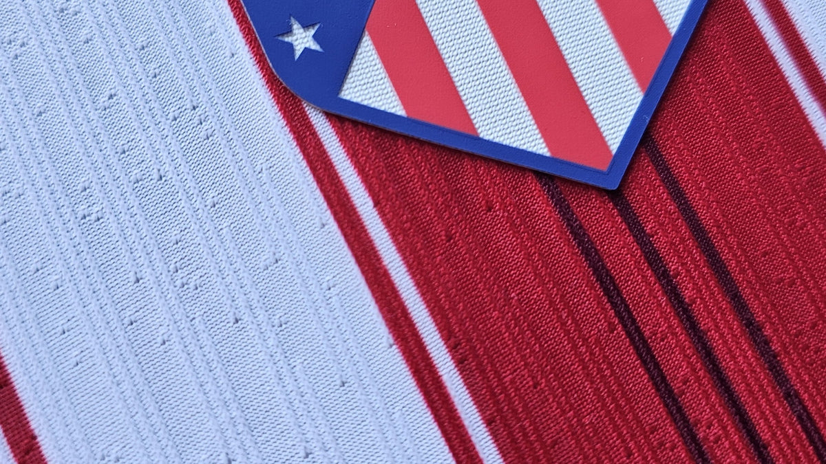 Close-up of an Atletico Madrid football shirt from the fanshop match tier. A heat-applied crest sits raised on the surface and the white stripe is a perforated engineered knit, the construction details that separate the tiers.