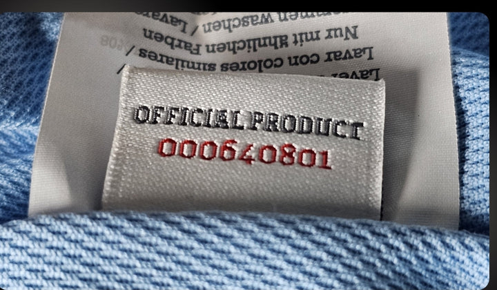 Close-up of a white woven label inside a light blue football shirt, showing the words Official Product above a red stitched serial number.