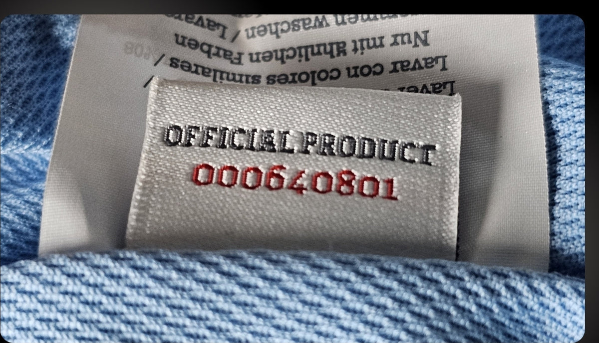 Close-up of a white woven label inside a light blue football shirt, showing the words Official Product above a red stitched serial number.