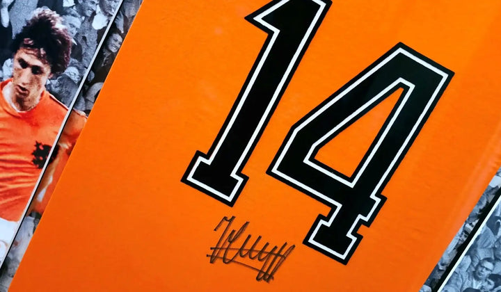 Signed orange Netherlands 1974 football shirt with black number 14, framed behind glass with printed Johan Cruyff photographs beside it.