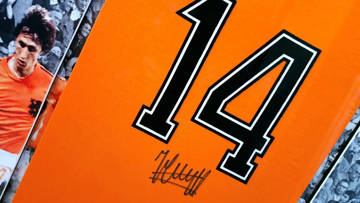 Signed orange Netherlands 1974 football shirt with black number 14, framed behind glass with printed Johan Cruyff photographs beside it.