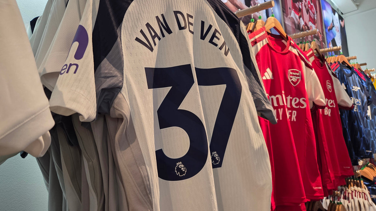 Row of football shirts on a rail, a white Tottenham away shirt named VAN DE VEN 37 in the foreground, red Arsenal shirts behind.