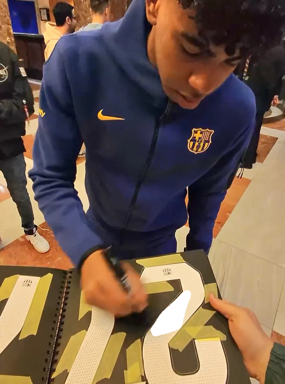 Lamine Yamal signing the No. 27 on a football shirt with a black pen. Signature blocked with white drawing.