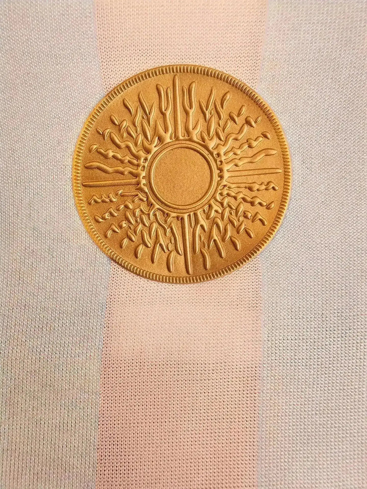 Crest detail of golden emblem with sun design on Argentina soccer jersey striped in blue and white.