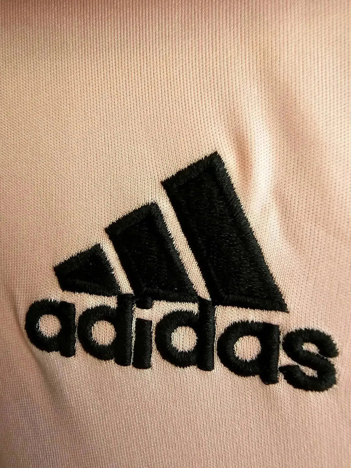 Close-up detail of the black embroidered adidas wordmark on a light pink soccer jersey.