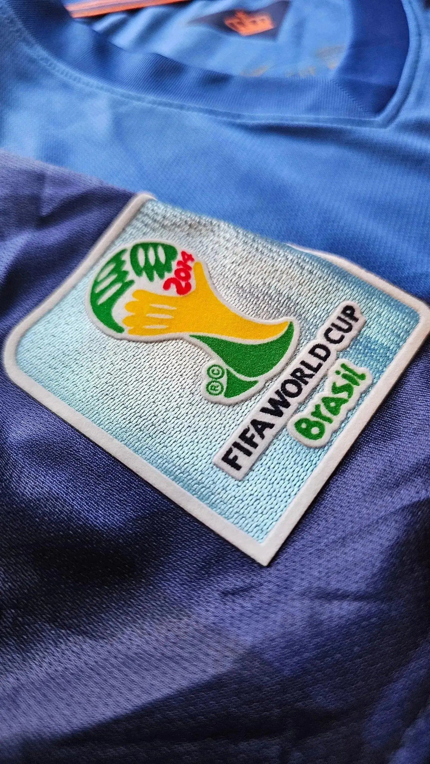 Crest detail featuring FIFA World Cup logo with '2014' and 'Brasil' on blue and orange football shirt.