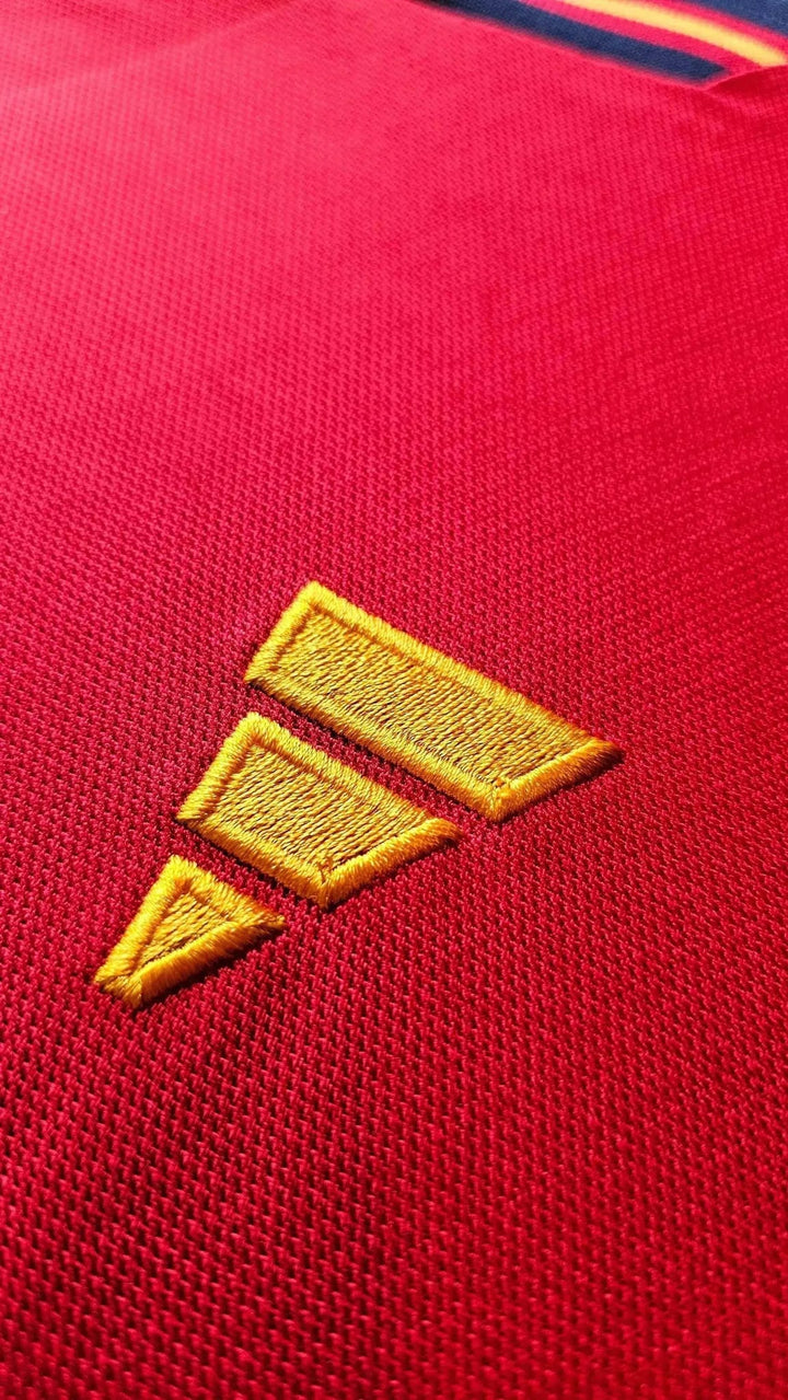 Close-up of the gold adidas three-stripe logo embroidered on Lamine Yamal's red Spain shirt, with the navy-and-gold sleeve trim visible.