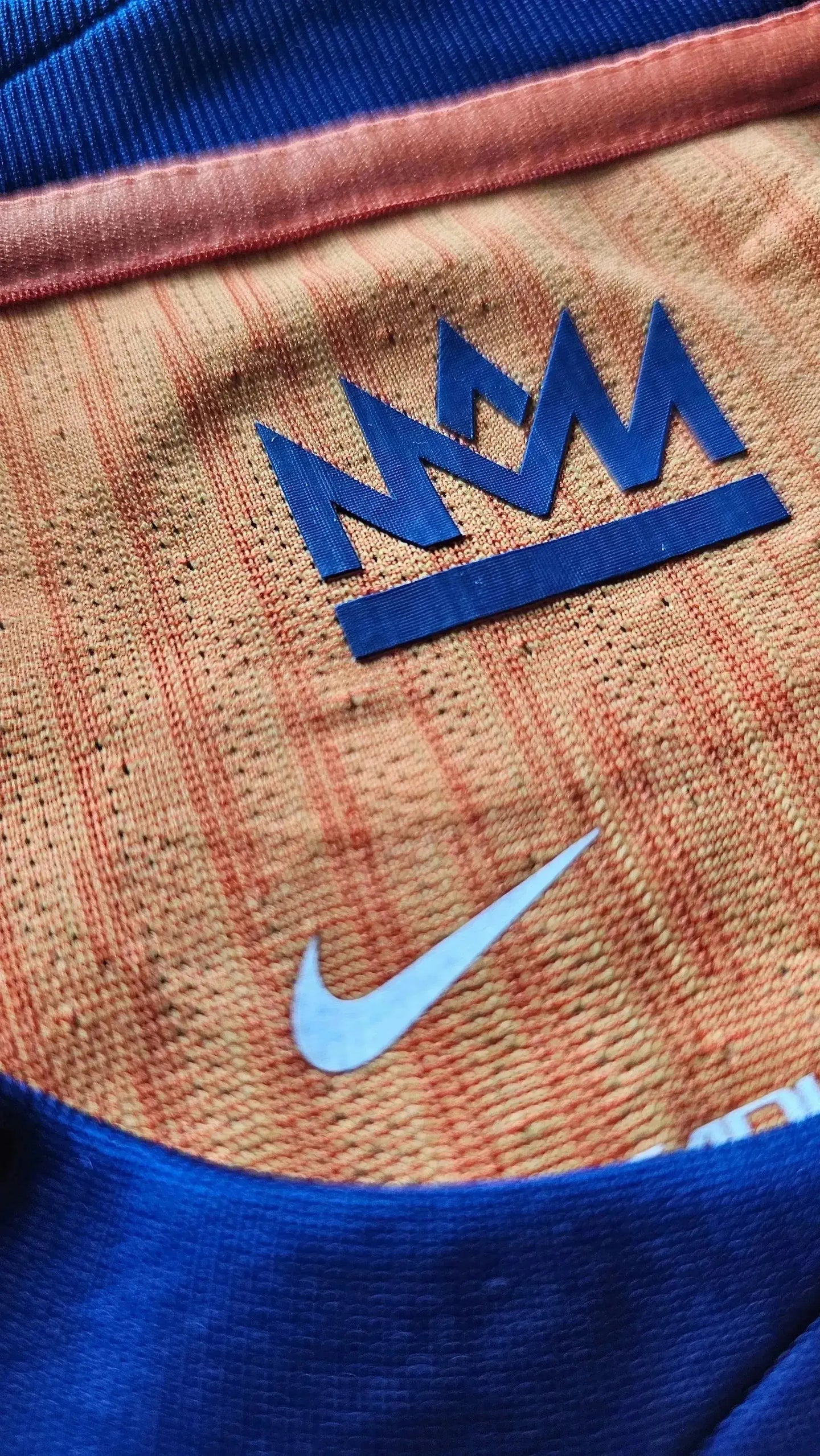 Close-up of orange shirt featuring Nike logo and crest with a crown detail.