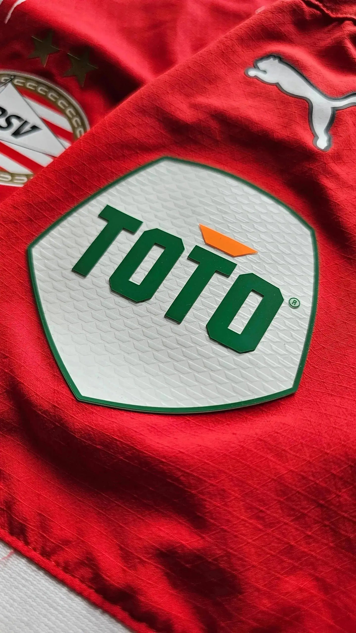 Crest detail showing PSV Eindhoven badge with two stars and TOTO sponsor logo on red fabric.