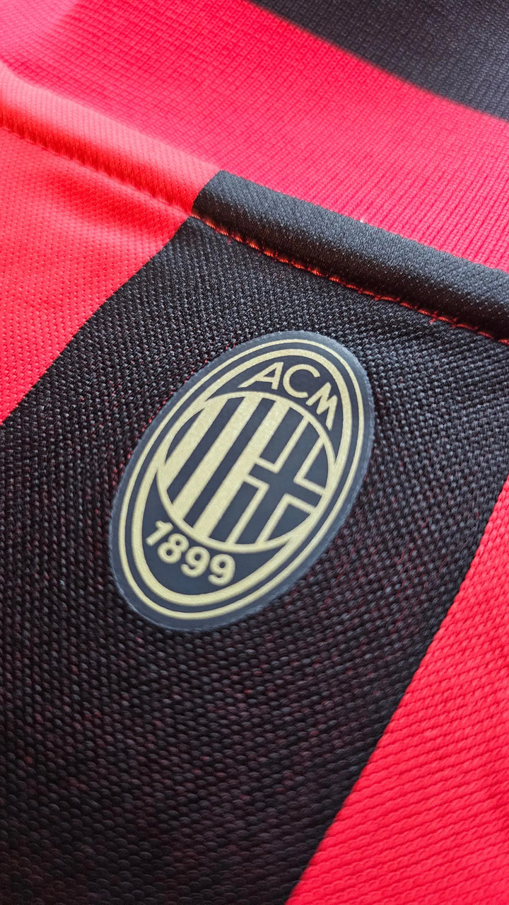 Close-up of AC Milan crest with gold details, featuring the year 1899 on a red and black striped background.