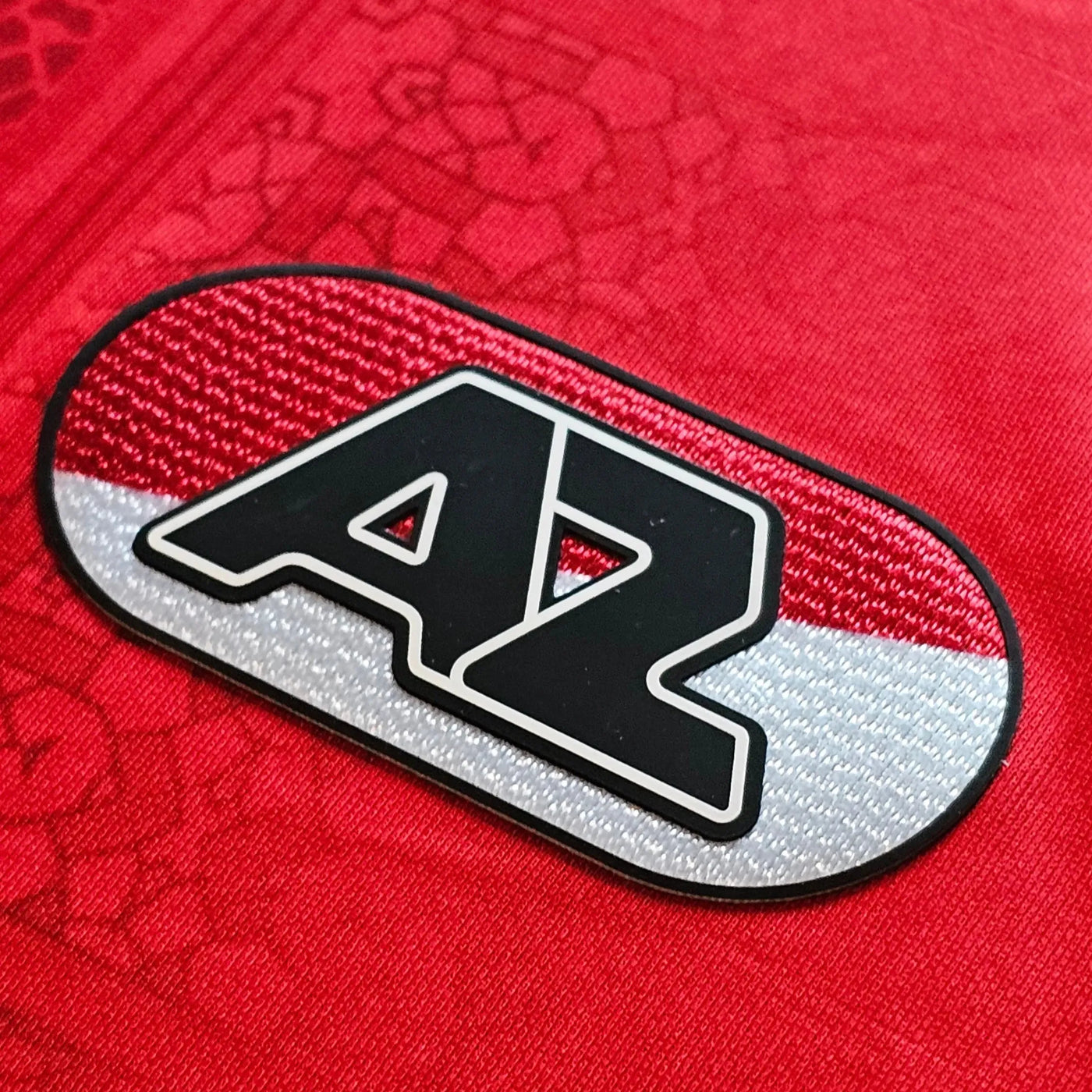 AZ Alkmaar crest on a red football shirt, oval red and silver patch with bold black AZ letters and fine woven texture.
