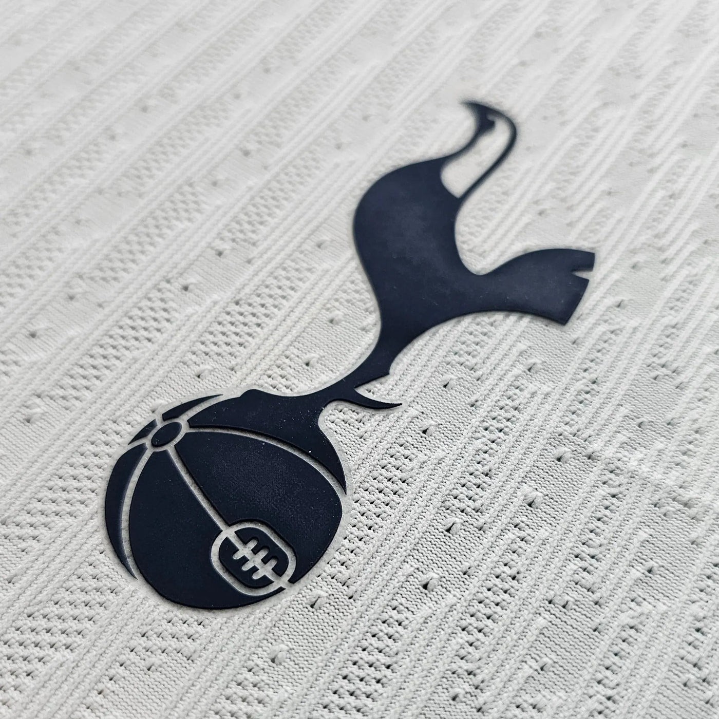 Tottenham Hotspur cockerel emblem in navy on a white football shirt with textured knit rows and small perforations, matte rubber finish.