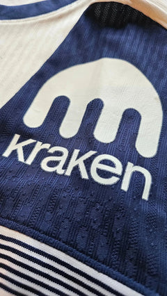 Close-up of Kraken sponsor logo on a blue and white football shirt.