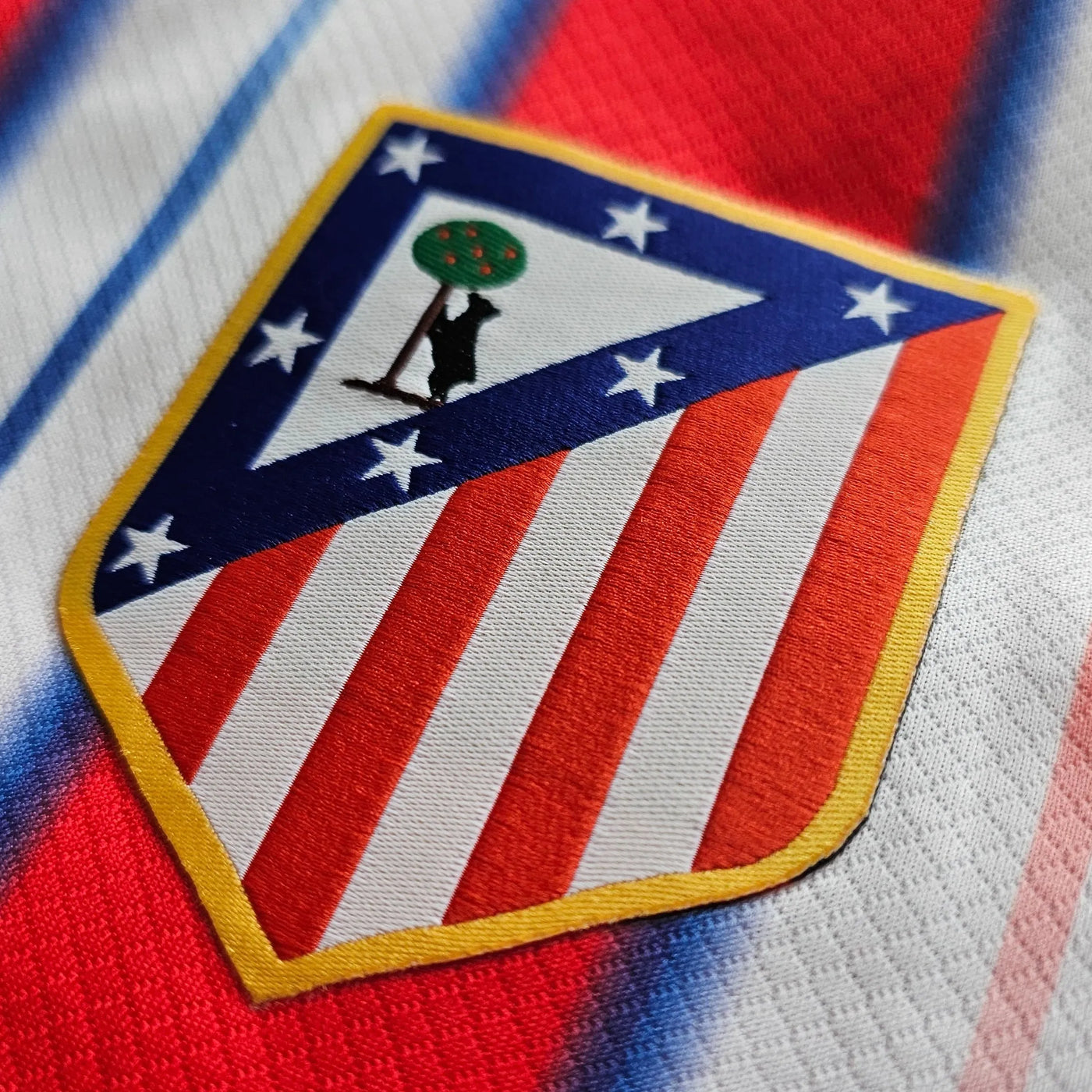 Atlético de Madrid crest on a white football shirt with red and blue gradient stripes, a shield with red and white stripes, a blue border with white stars and a yellow edge.