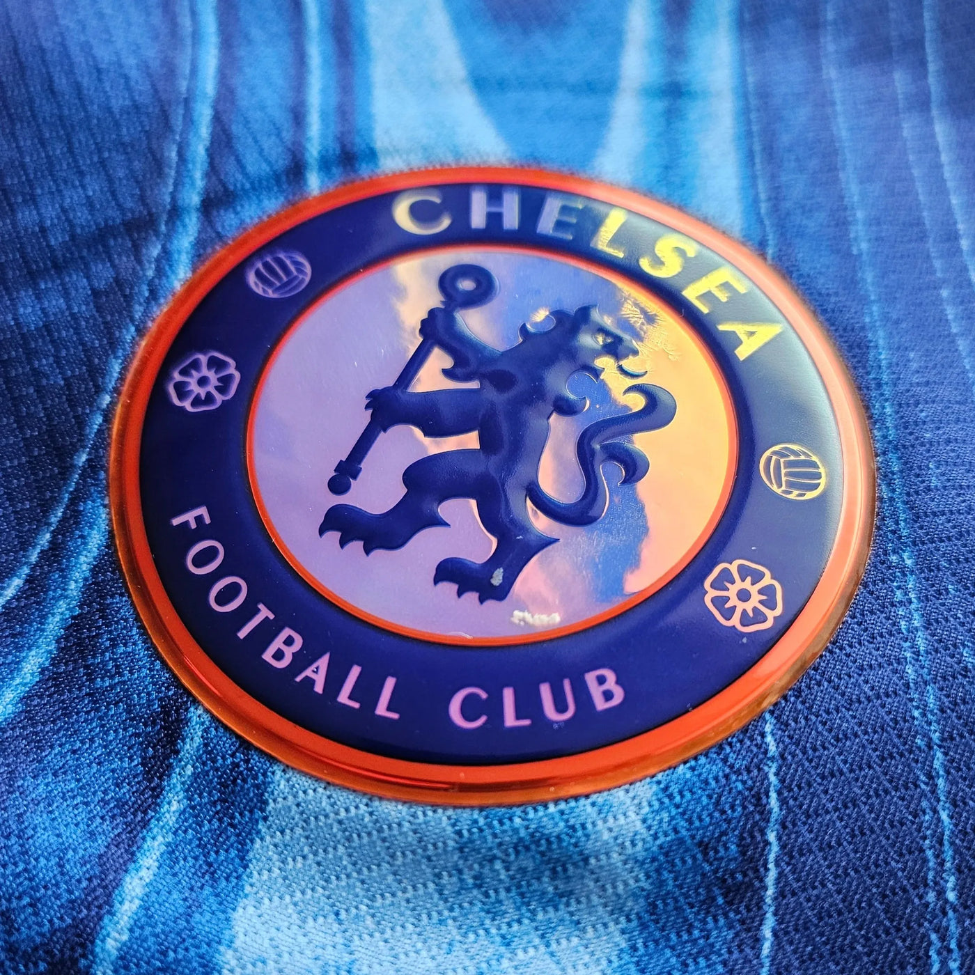 Chelsea Football Club circular crest on a blue football shirt with wavy tonal stripes, a navy roundel with an orange rim and a raised rubber finish showing the lion.
