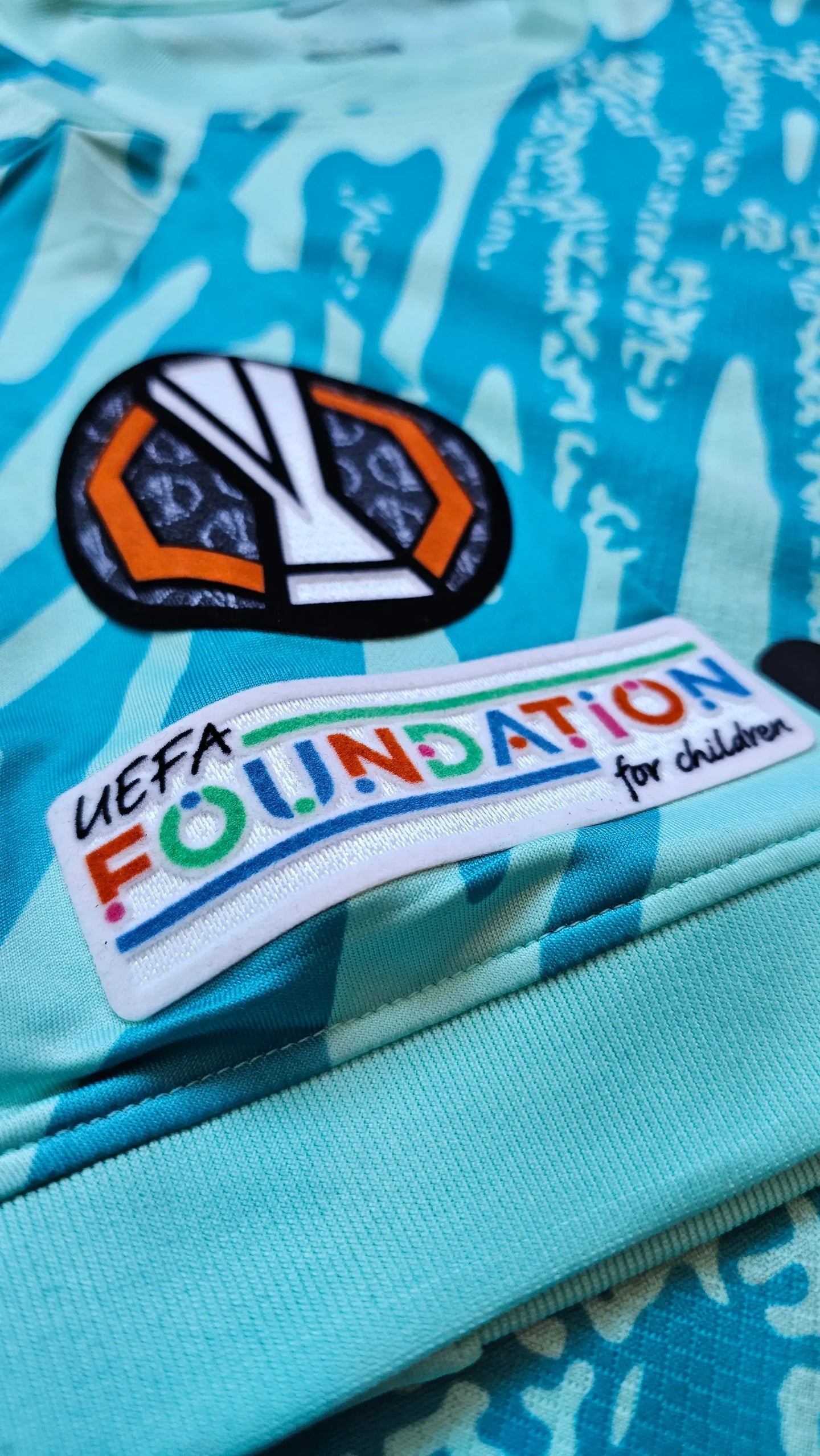 Close-up of UEFA Foundation patch and competition badge on a blue football shirt.