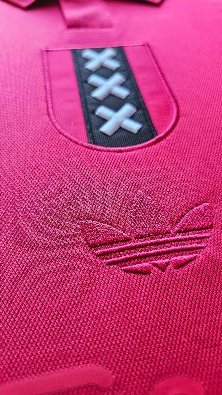 Detail of Ajax crest featuring three X's and adidas logo on a pink football shirt.