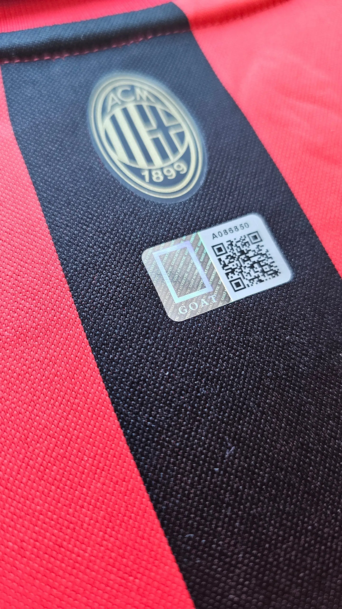 Close-up of the AC Milan crest and authenticity tag with QR code on a red and black football shirt.
