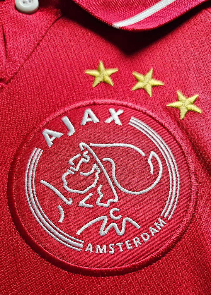 Crest detail of Ajax with three stars and 'AJAX AMSTERDAM' in white on a red background.