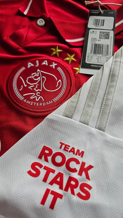 Front view of Ajax football shirt featuring red color, crest with two stars, and 'TEAM ROCK STARS IT' text.