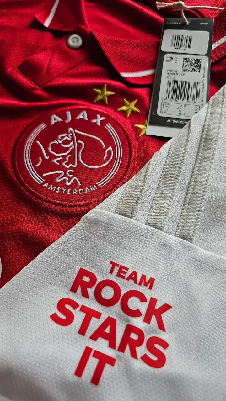 Front view of Ajax football shirt featuring red color, crest with two stars, and 'TEAM ROCK STARS IT' text.