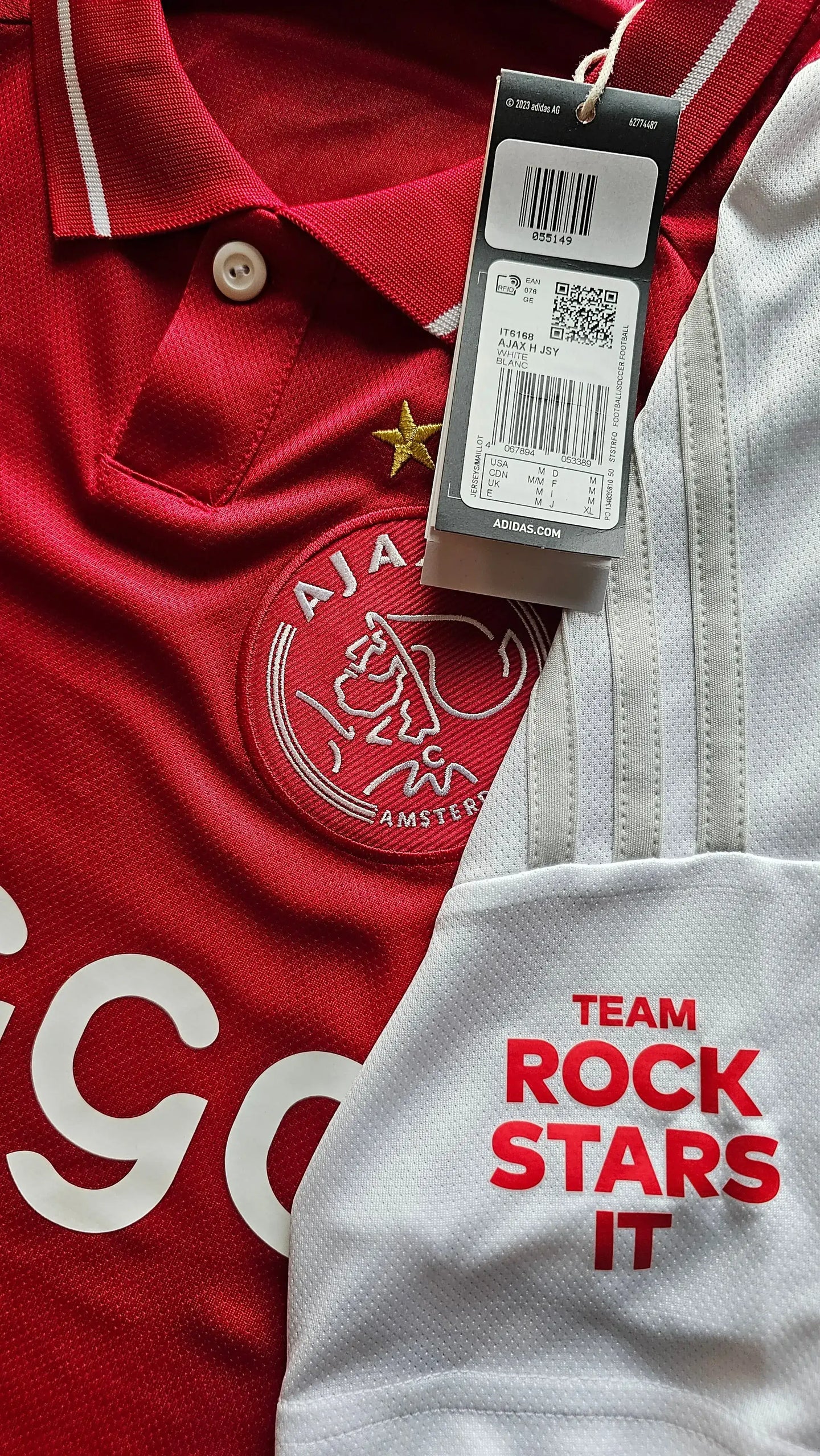 Front view of red Ajax football shirt with white details and embroidered Ajax crest. Tags visible.
