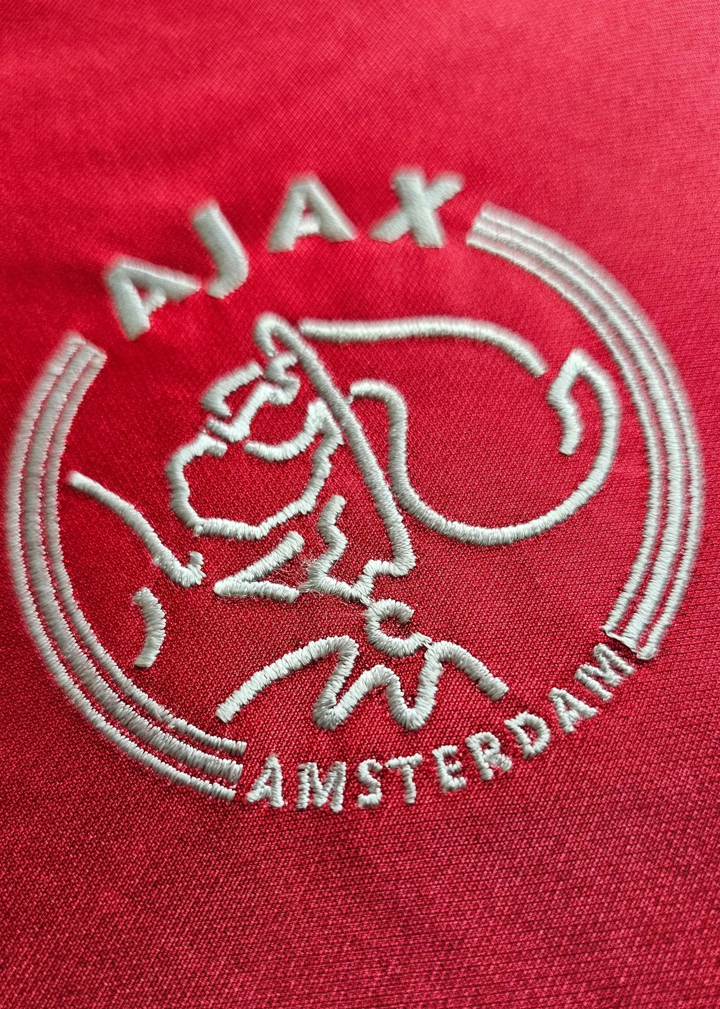 Crest detail of AFC Ajax featuring embroidered logo and 'AMSTERDAM' text on a red background.