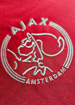 Crest detail of AFC Ajax featuring embroidered logo and 'AMSTERDAM' text on a red background.