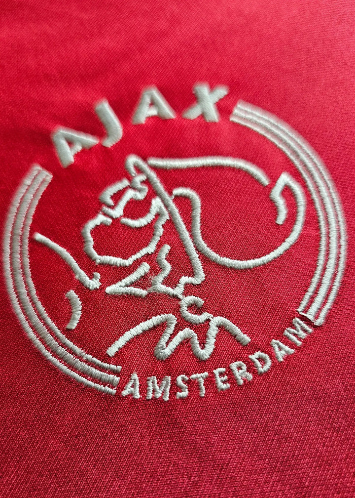 Crest detail of AFC Ajax featuring embroidered logo and 'AMSTERDAM' text on a red background.