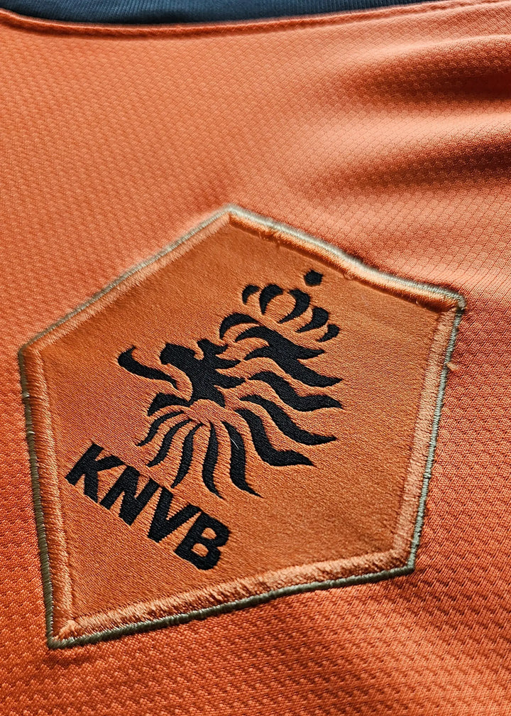 Crest detail of the Holland football shirt featuring the KNVB lion emblem in black on an orange background.