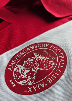 Crest detail showing AFC Ajax logo with text 'AMSTERDAMSCHE FOOTBALL CLUB' and dates '1900-2025'.