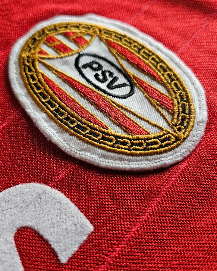 Close-up of the PSV Eindhoven crest on a red football shirt with intricate embroidery.