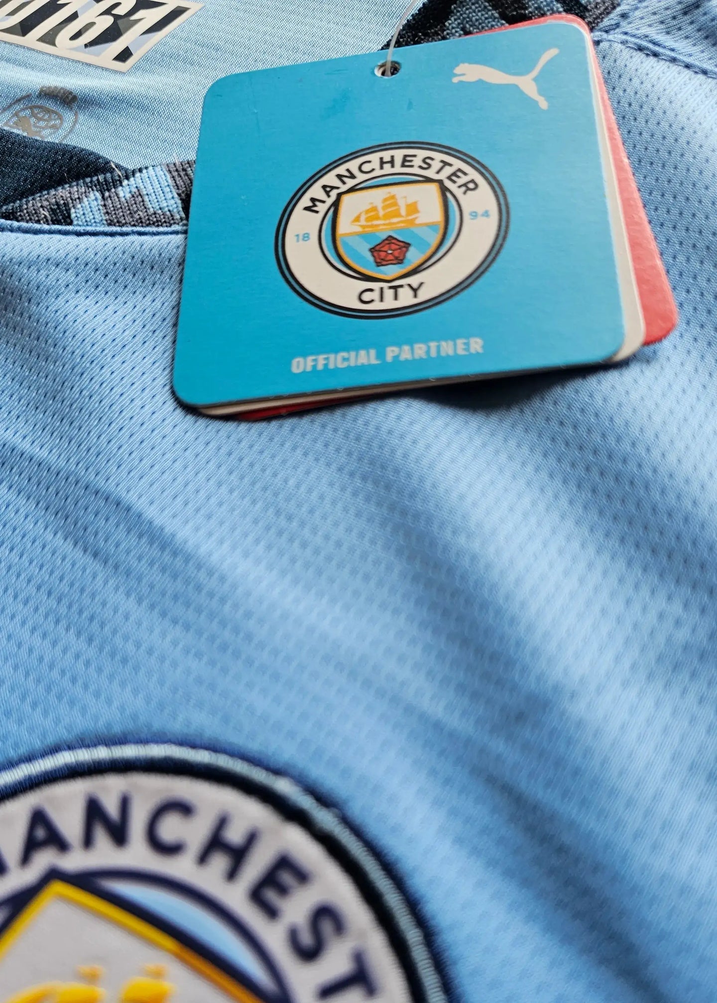 Close-up of Manchester City crest on blue football shirt with 'OFFICIAL PARTNER' tag visible.
