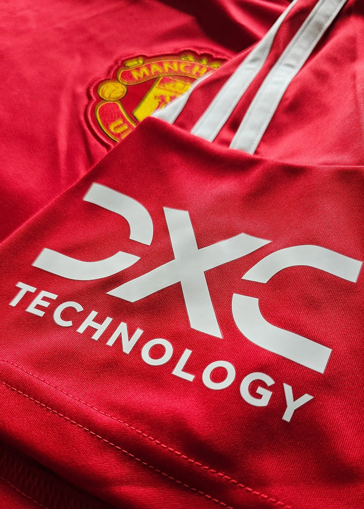 Detail view of Manchester United crest and CX TECHNOLOGY logo on a red football shirt.