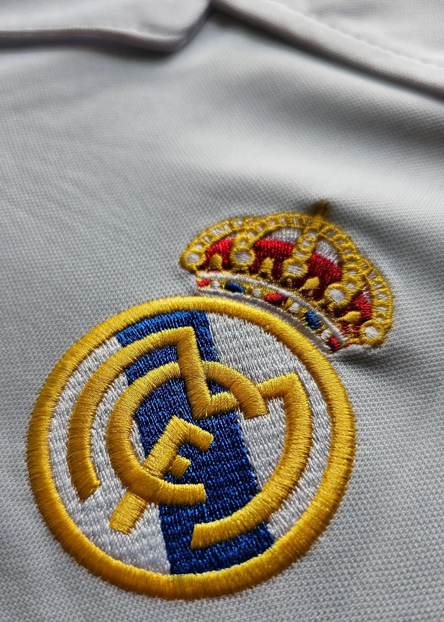 Detailed view of Real Madrid CF crest with gold crown and blue stripe on a white football shirt.