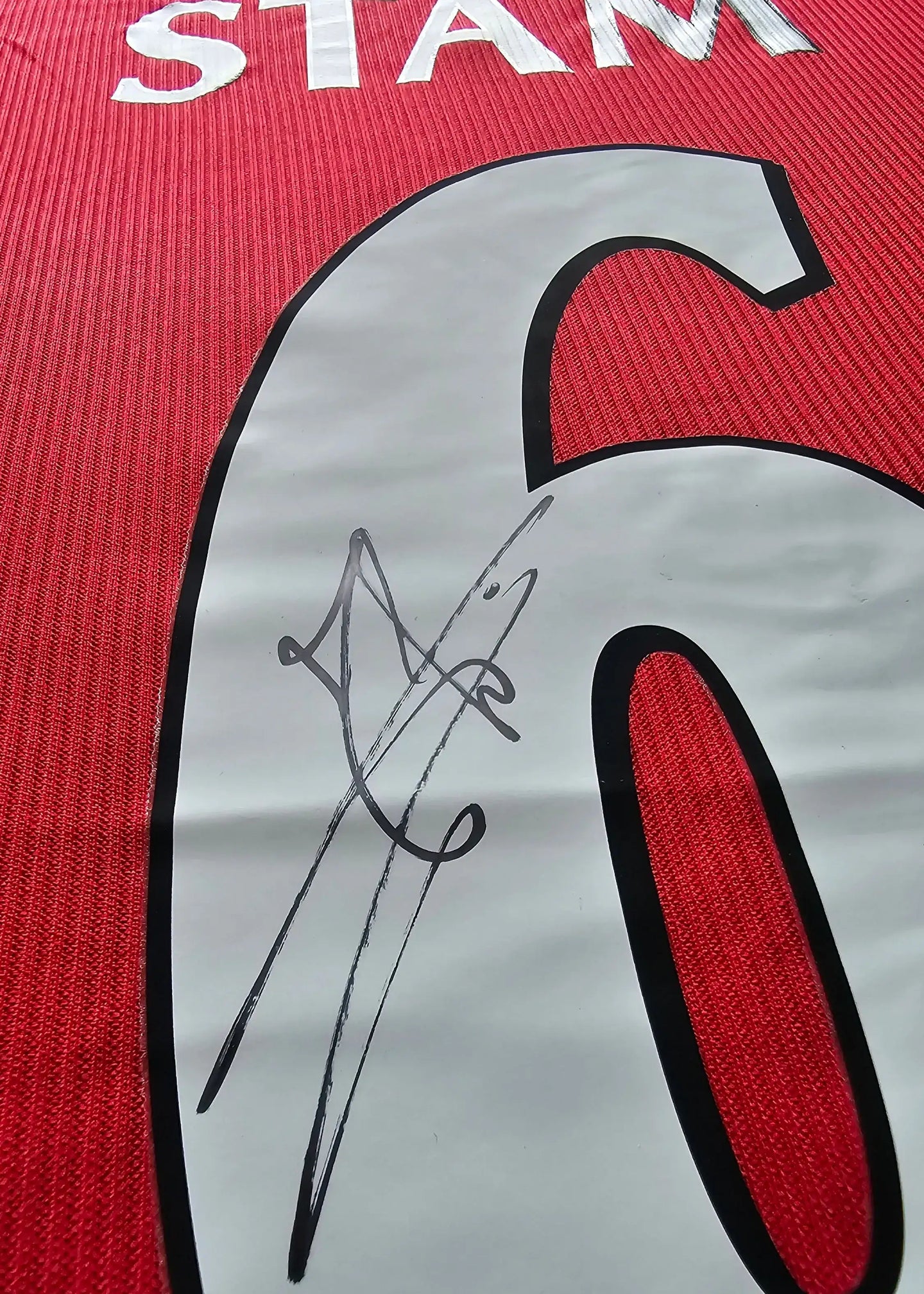 Detail showing the signature of Stam on the No. 6 of a red Manchester United soccer jersey.