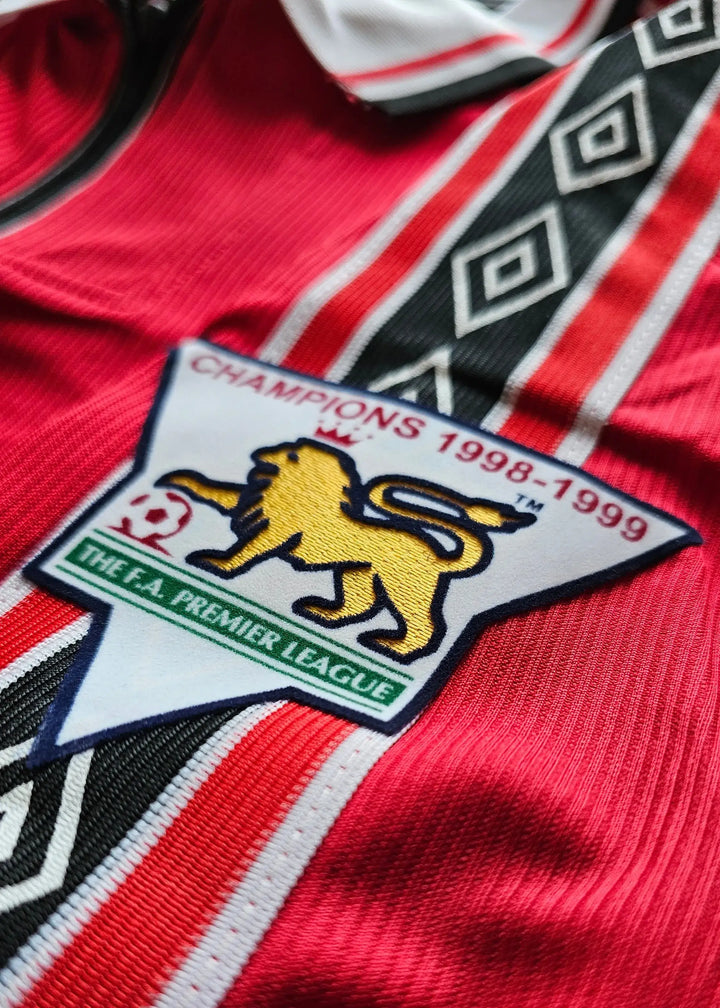 Detail showing Manchester United crest with the text CHAMPIONS 1998-1999 and F.A. PREMIER LEAGUE.