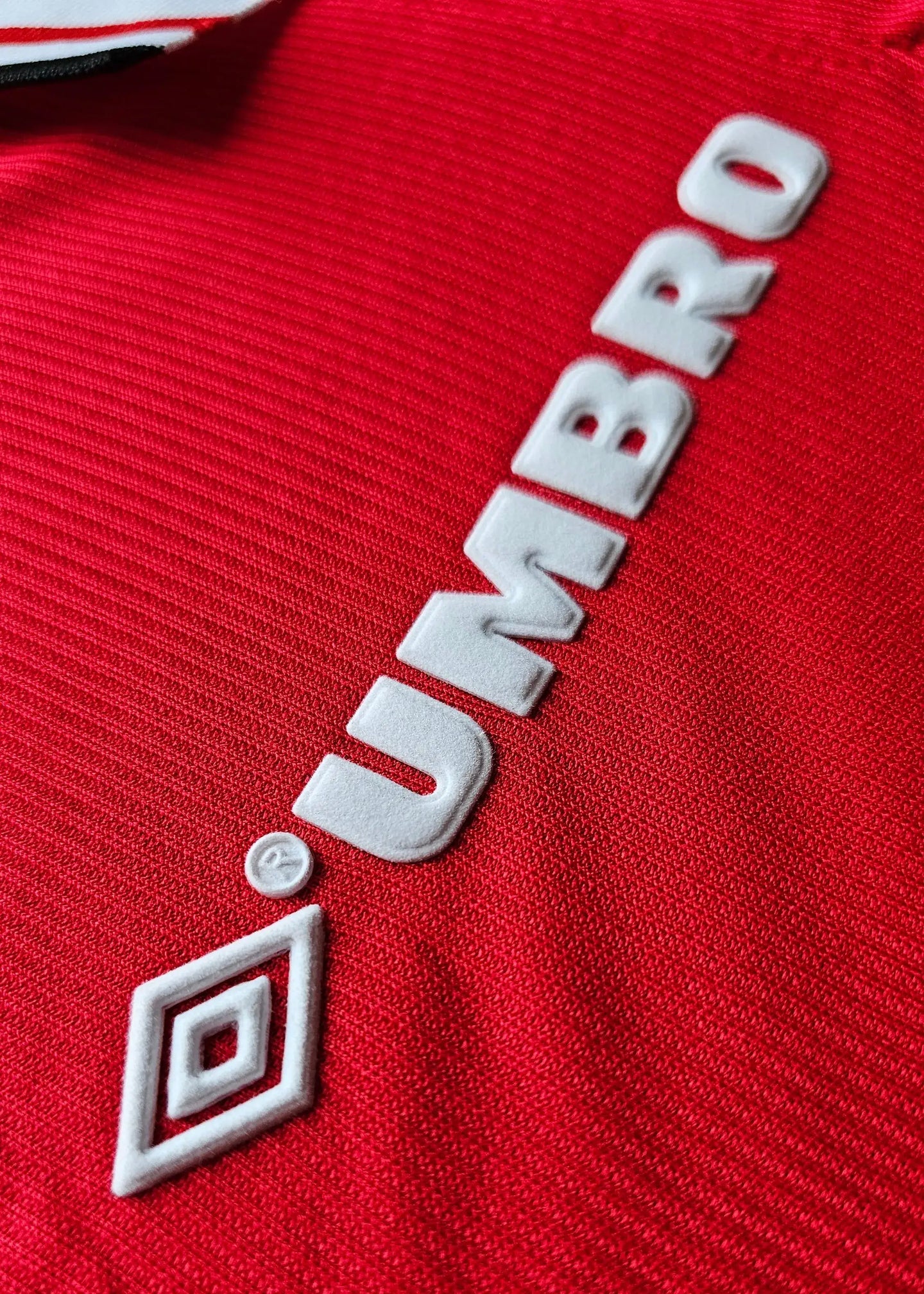 Close-up of red fabric with white 'UMBRO' logo detail on the signed football shirt.