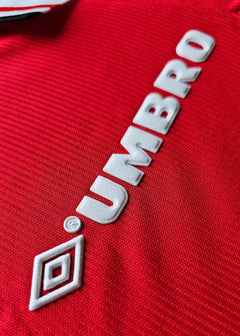 Close-up of red fabric with white 'UMBRO' logo detail on the signed football shirt.