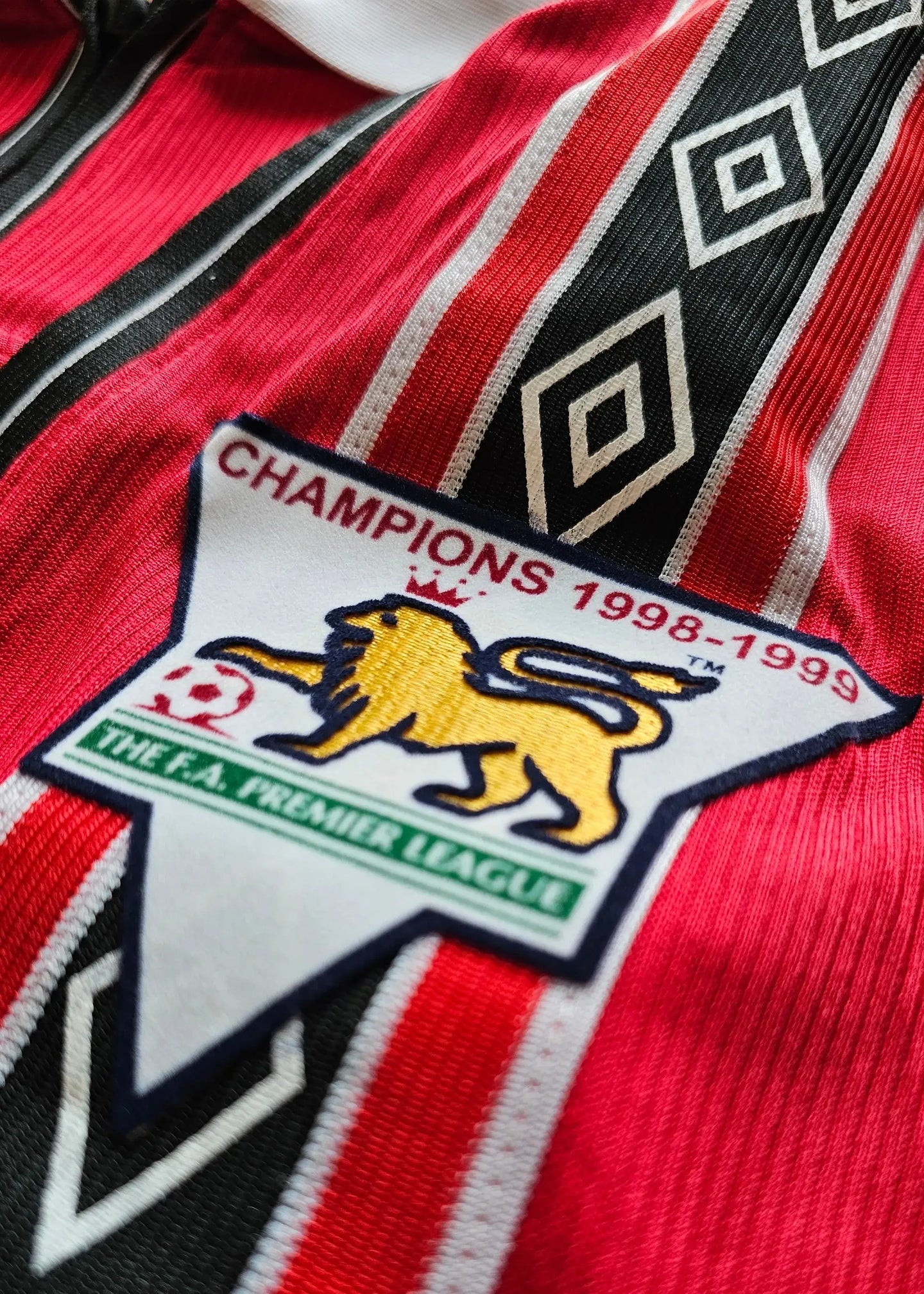 Crest detail featuring lion emblem with text 'CHAMPIONS 1998-1999' and 'THE F.A. PREMIER LEAGUE'.