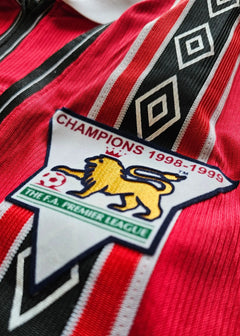 Crest detail featuring lion emblem with text 'CHAMPIONS 1998-1999' and 'THE F.A. PREMIER LEAGUE'.