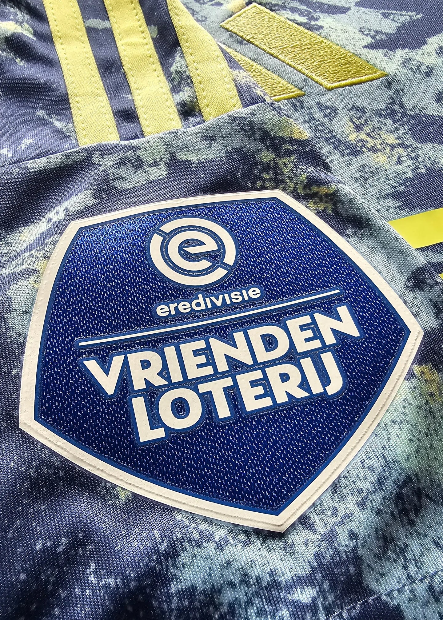 Close-up of Vrienden Loterij patch on Ajax shirt with yellow stripes and blue background.