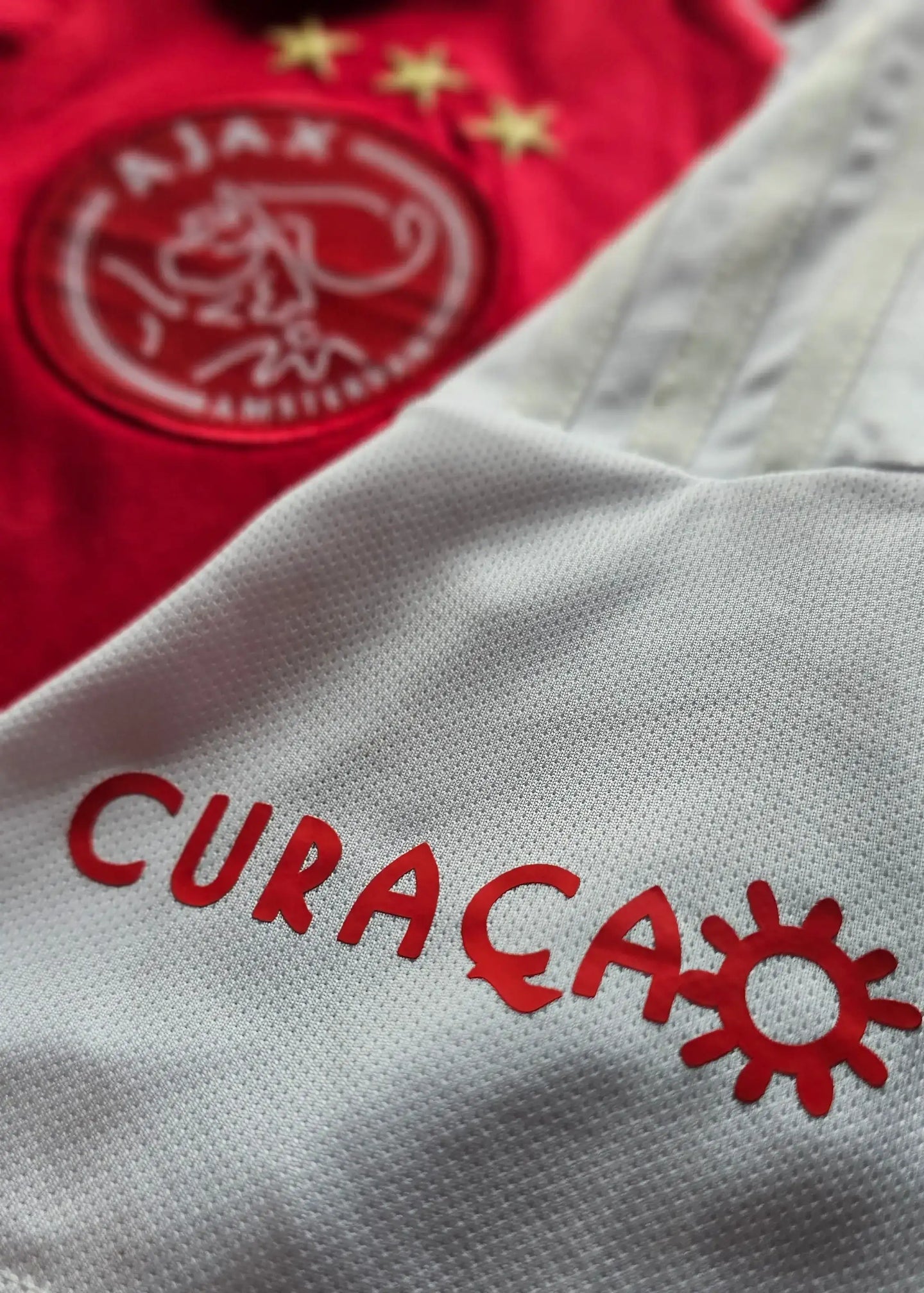 Close-up of Ajax crest with three stars and "CURACAO" printed in red on white fabric.