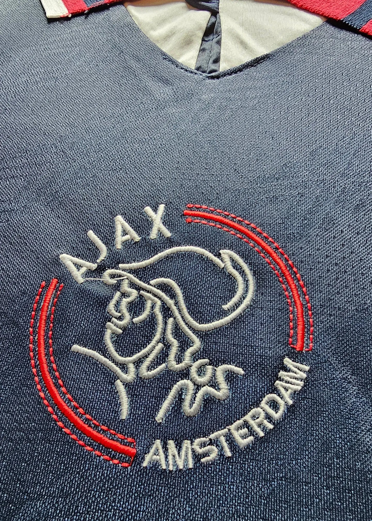 Embroidered crest of Ajax with 'AJAX' and 'AMSTERDAM' text, featuring red and white detailing.