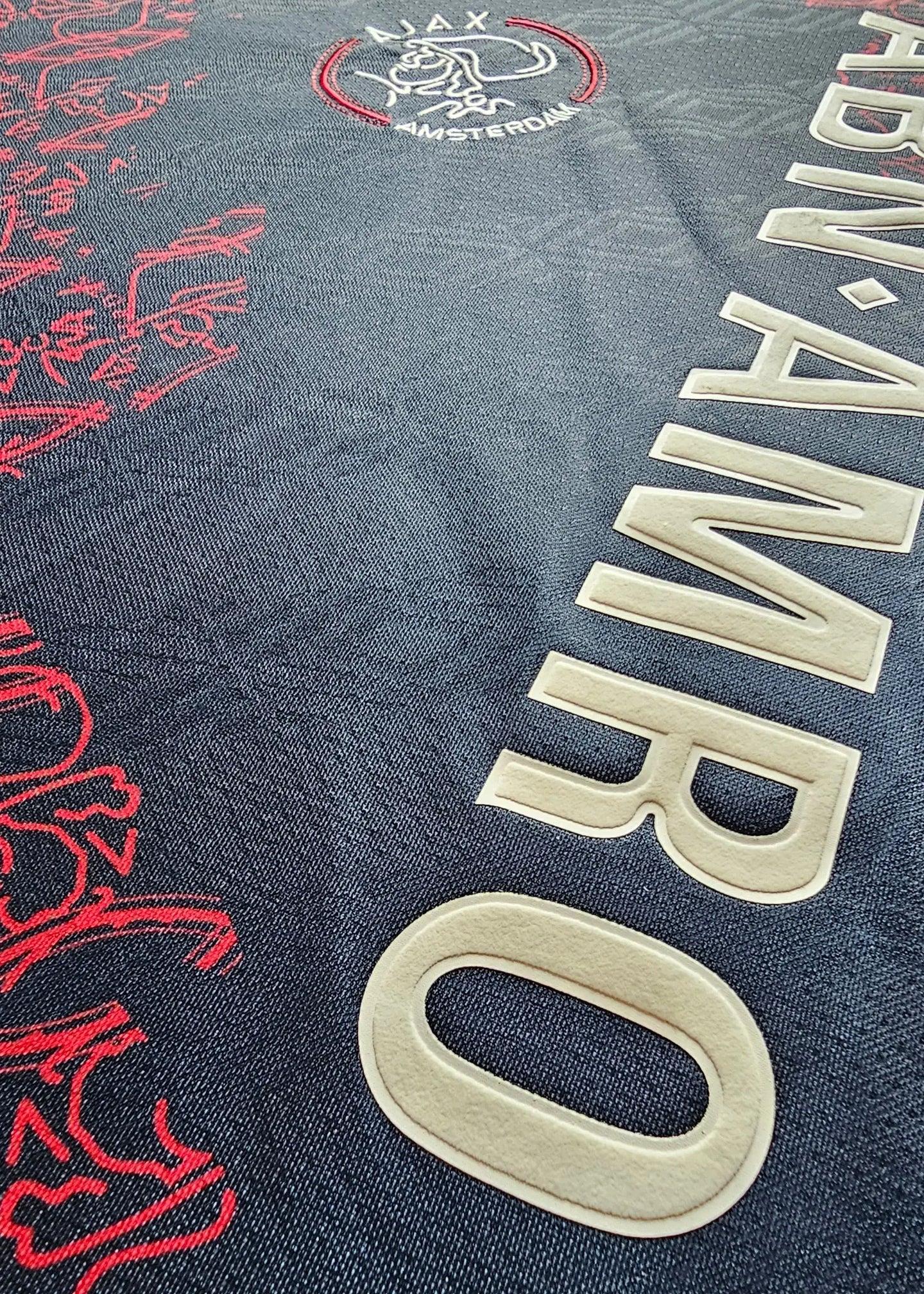 Crest detail featuring Ajax logo, red design elements, and the word 'AMSTERDAM' in prominent lettering.