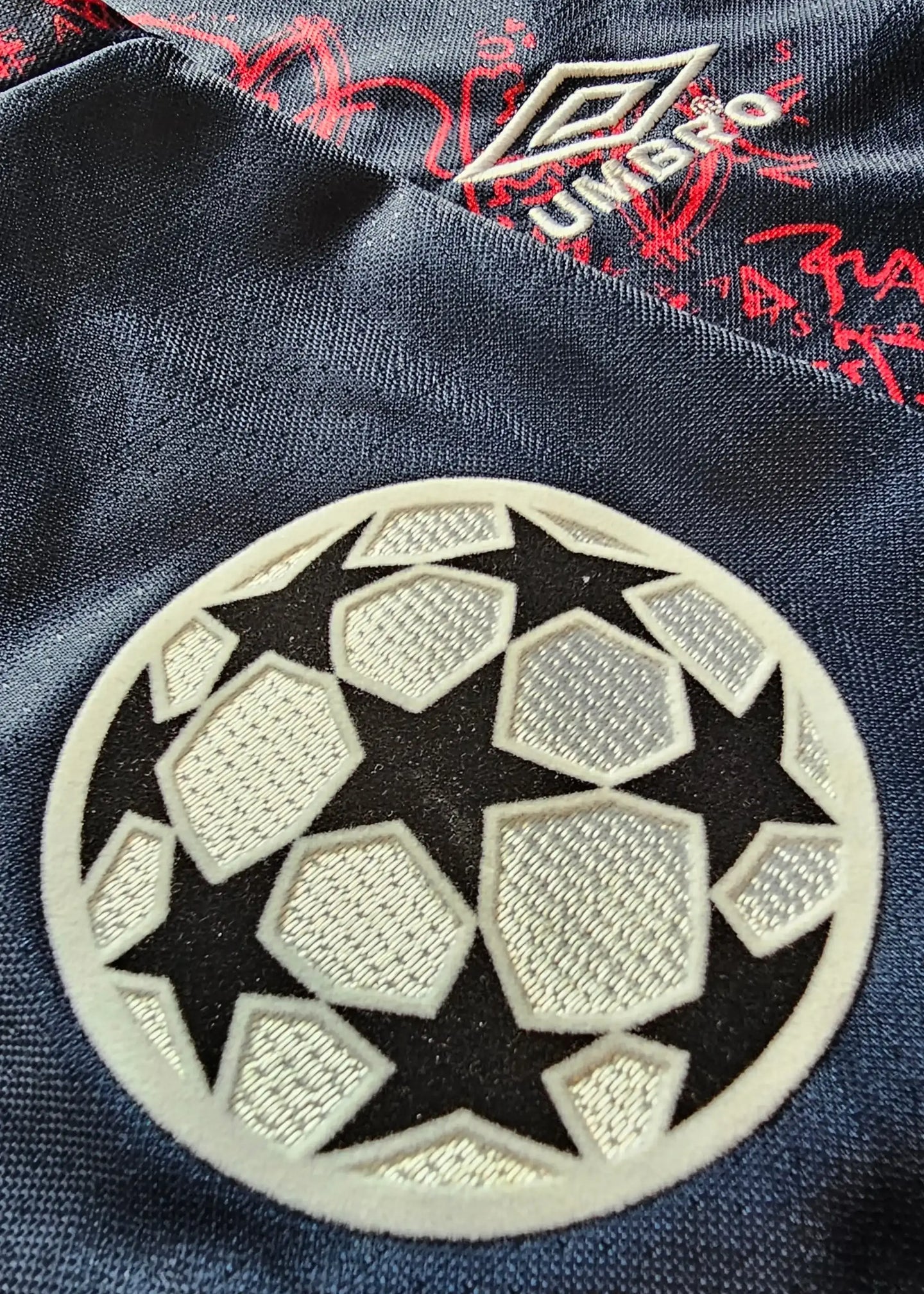 Crest detail showing the Champions League logo on a navy football shirt with red accents.