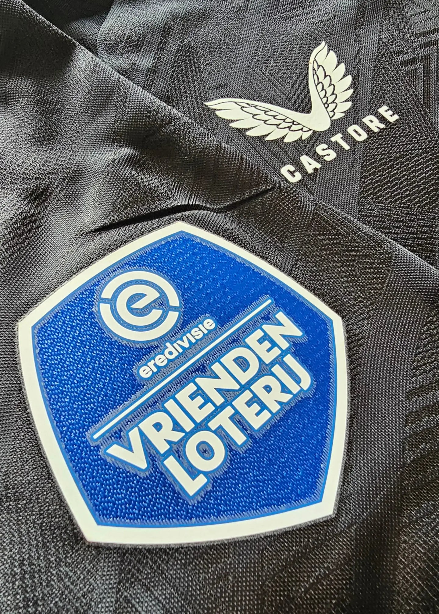 Close-up showing the blue 'VRIENDEN LOTERIJ' patch and 'Castore' logo on a black football shirt.