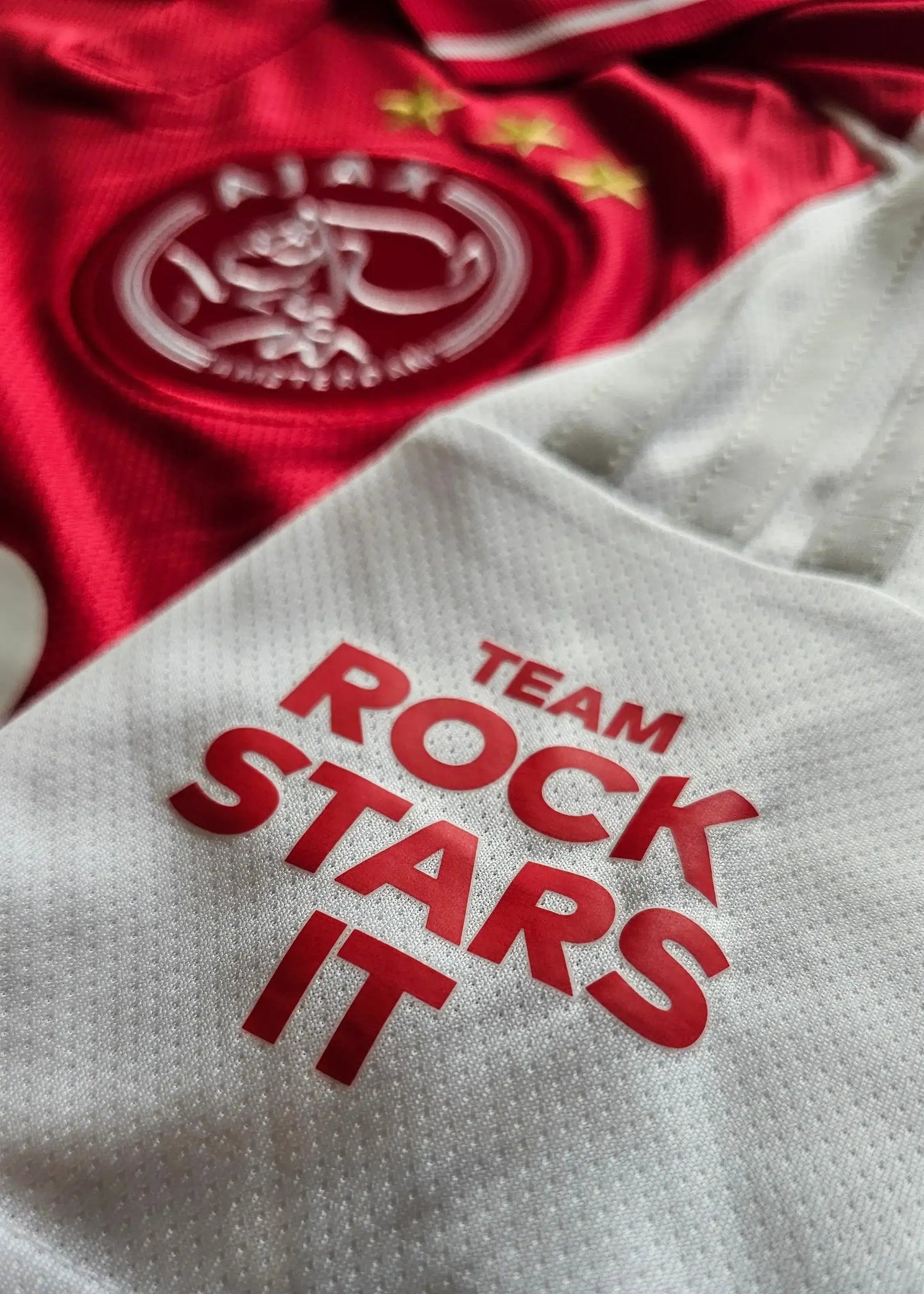 Ajax crest with two stars above and text 'TEAM ROCK STARS IT' in red on white fabric.