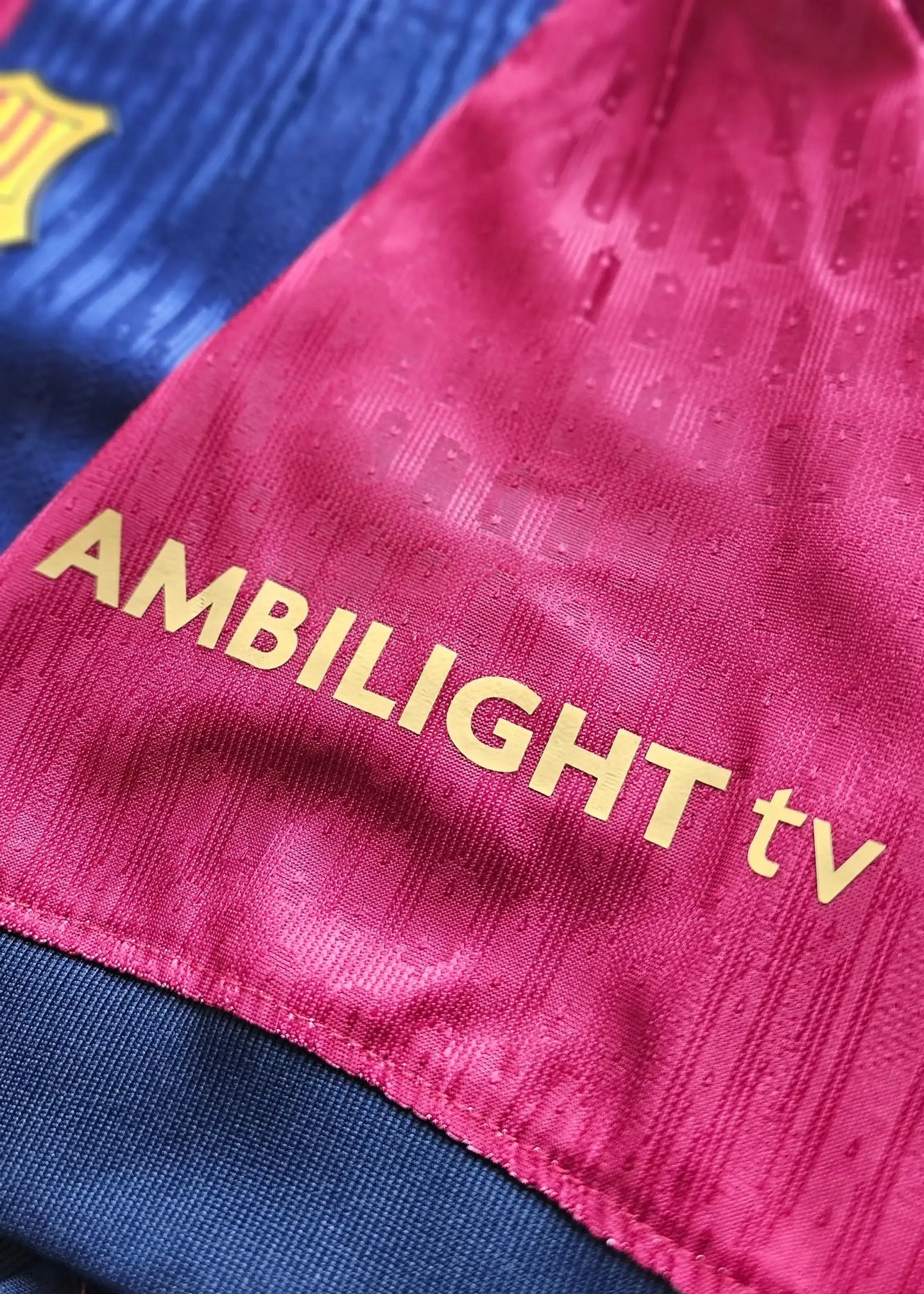 Crest detail showing the AMBILIGHT tv sponsor in gold on the pink part of the Barcelona football shirt.